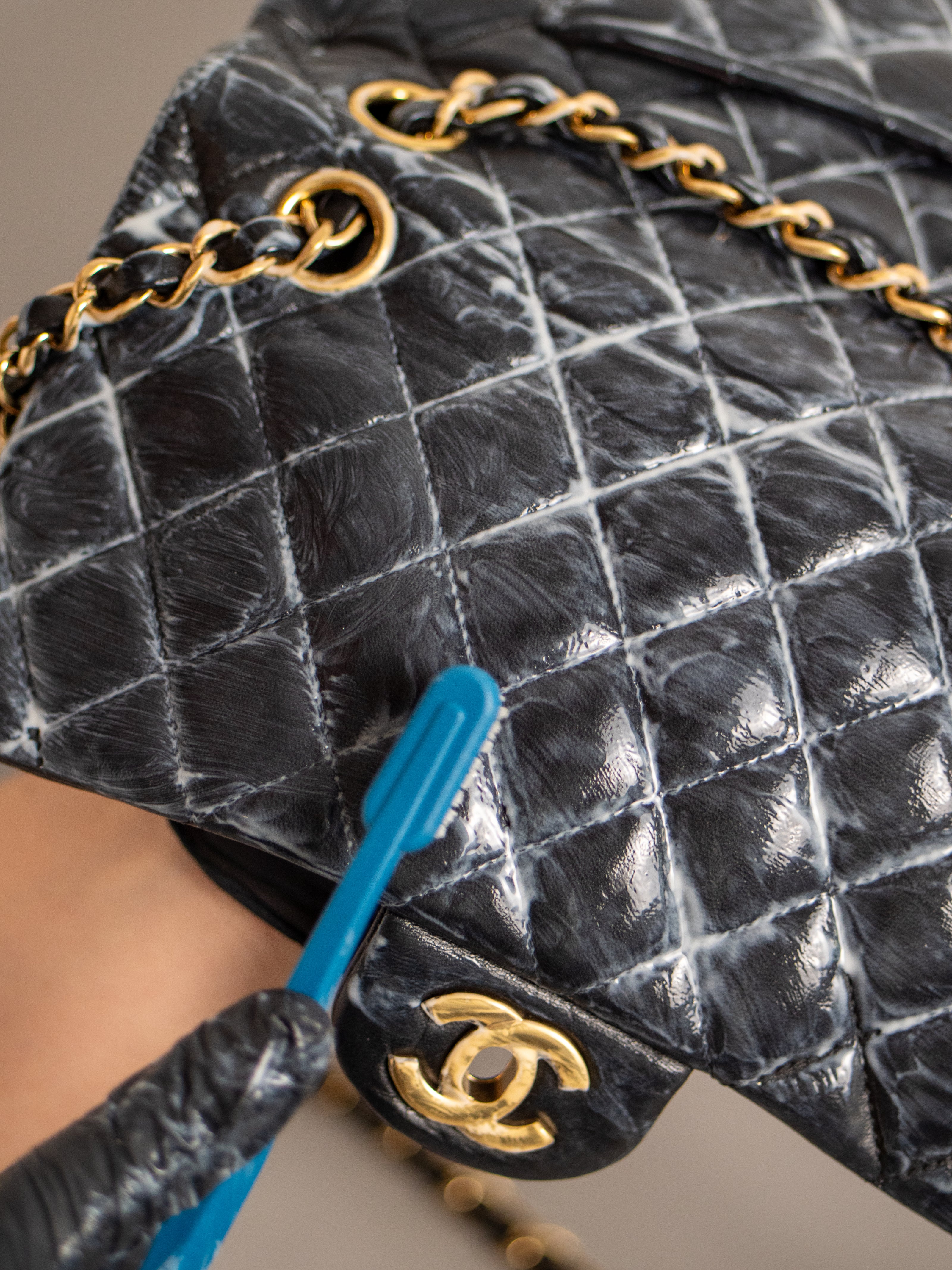 Black quilted handbag with gold chain and clasp, being cleaned with a blue brush.