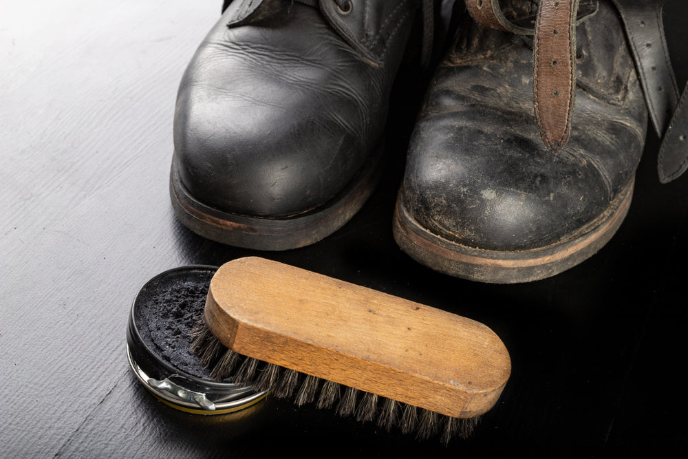 before and after comparison of a pair of leather black shoes side by side and leather wax and a brush on a black table