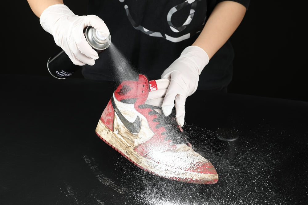 a man with white gloves spraying a white soap from an aerosol can onto a dirty red nike high top sneaker on black table