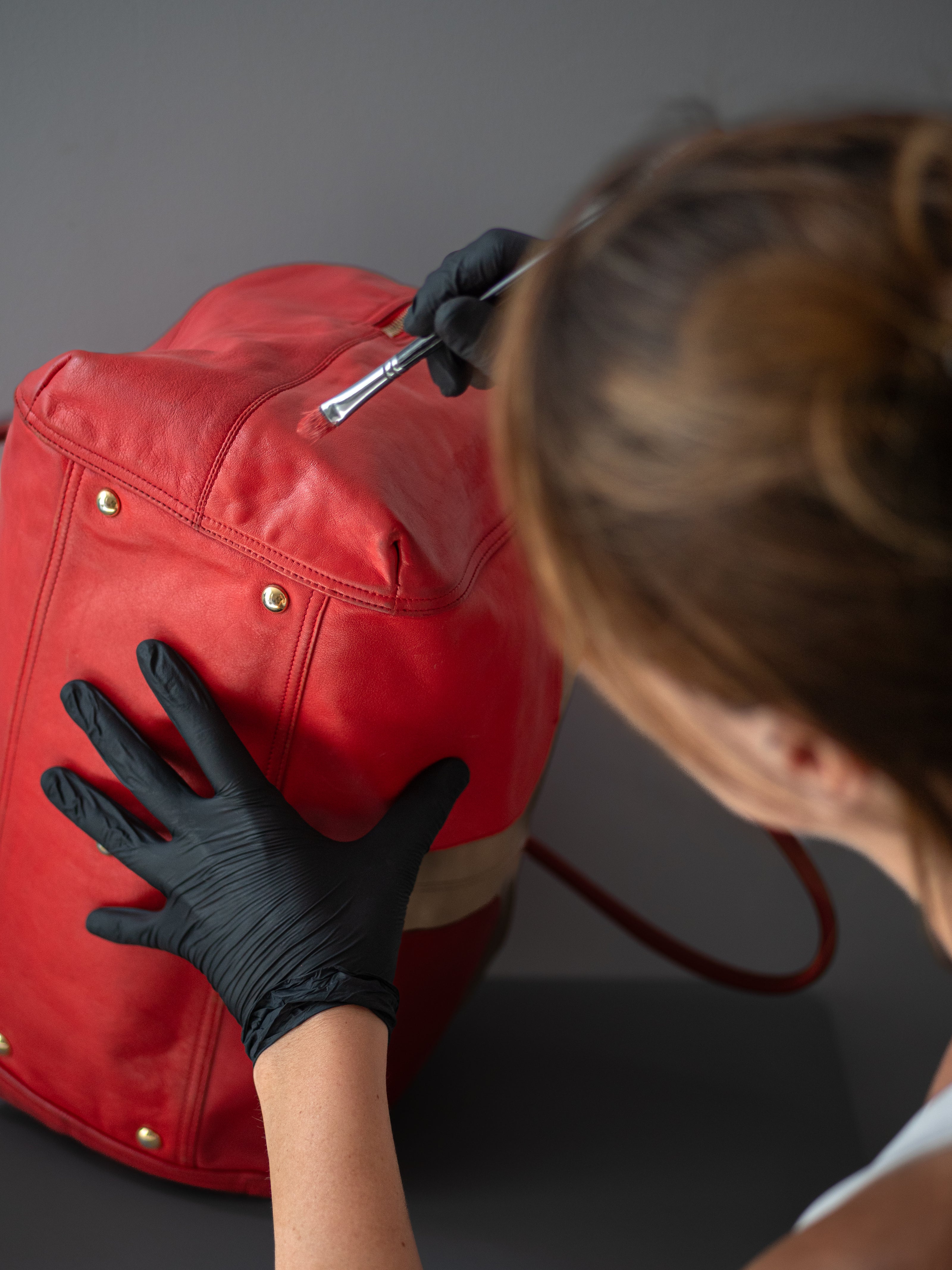 Person wearing black gloves working on a red leather bag against a gray background